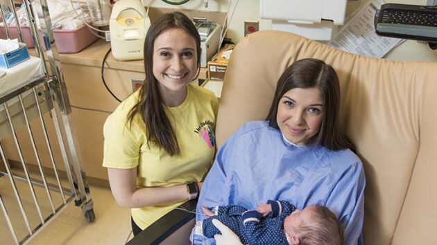 一位黄色衬衫的奥尔巴尼医疗中心工作人员站在一名妇女的左侧，在医院的床上抱着新生婴儿。